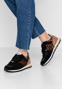 Svarta sneakers med leopardmönster, i mocka och tyg, med gröna detaljer och en vit sula. Bärs med uppvikta blå jeans.