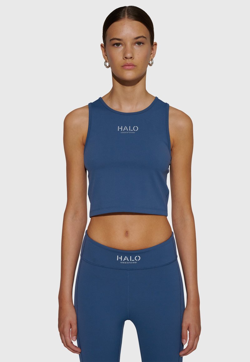 Ung kvinna som bär blå ärmlös crop-top och matchande högmidjade leggings med "HALO"-logotyp, stående mot en enfärgad ljus bakgrund.