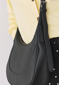 Sac à bandoulière en cuir noir texturé avec une sangle ajustable porté par-dessus un cardigan jaune et une jupe noire à pois.