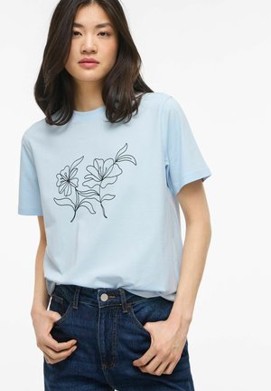 Joven con camiseta azul claro con diseño floral negro simple, metida dentro de unos vaqueros azul oscuro, de pie con las manos en los bolsillos.