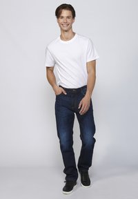Witte T-shirt en donkerblauwe jeans, beide in een casual pasvorm. Model staat met één hand in de zak, draagt zwarte sneakers. Eenvoudige achtergrond.