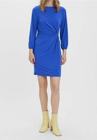 Modell klädd i en figurnära blå klänning med långa ärmar och en snurrdetalj vid midjan, tillsammans med gula ankelstövlar på en enfärgad bakgrund.