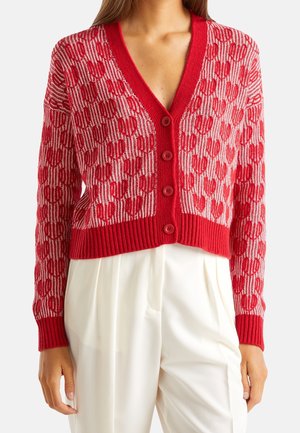Cardigan rouge boutonné avec un motif de cœurs roses porté par-dessus un pantalon blanc plissé, montré de la poitrine aux hanches.