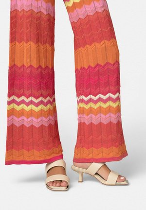 Beine, die mehrfarbige, zackige Muster-Hose mit weitem Bein tragen, kombiniert mit beigen offenen Sandalen mit Absatz, auf weißem Hintergrund.