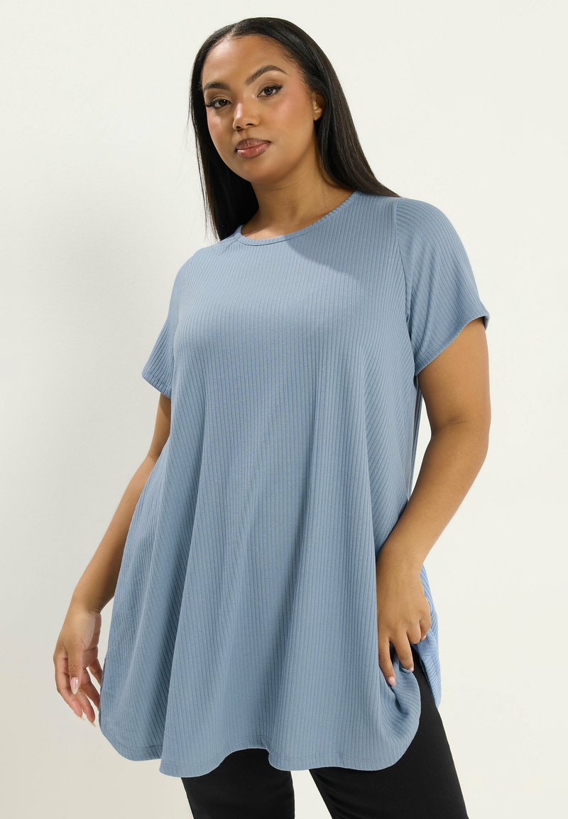 Donna che indossa una maglia azzurra chiara a maniche corte e a coste, dall'ampio taglio, abbinata a pantaloni neri, in piedi davanti a uno sfondo uniforme.