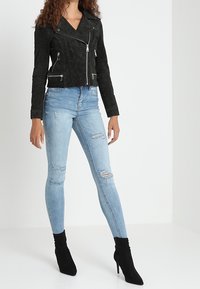 Sort ruskind motorcykeljakke med lynlåsdetaljer, parret med lyseblå slidte skinny jeans og sorte ankelstøvler. Lige silhuet.
