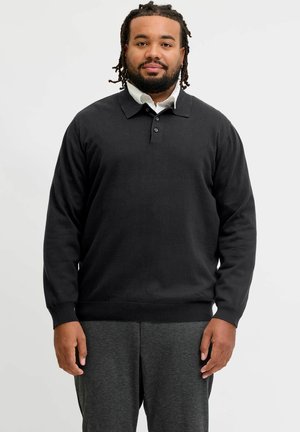 Mand med fletninger i håret og skæg iført en sort langærmet poloshirt over en hvid skjorte med krave og mørkegrå bukser, stående.