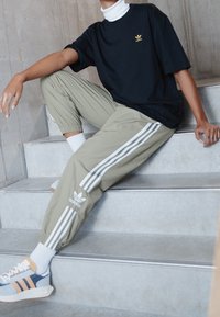 Person som sitter på betongtrappor iförd svart Adidas-tröja, beige Adidas träningsbyxor med vita ränder, vita strumpor och blå sneakers.