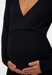 Czarna prążkowana sukienka ciążowa z dekoltem w serek, długimi rękawami i dopasowaną talią podkreślającą zaokrąglony brzuch, ręce umieszczone na brzuchu.