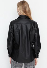 Camicia in pelle nera con colletto, maniche lunghe e trama sottile. Indossata con pantaloni a quadri grigi, con focus sulla vista posteriore.