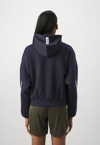 Marinblå kort hoodie med en bred huva och elastisk nederkant. Har en vit logotag på ryggen och avslappnade ärmar. Kombineras med olivgröna shorts.