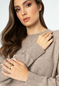 Hellbrauner Strickpullover mit gerippten Bündchen und einem gestickten Design. Das Model trägt mehrere goldene Ringe an beiden Händen.