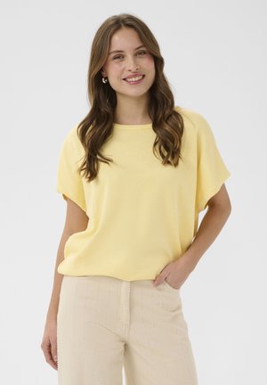 Kvinde med langt brunt hår iført en gul kortærmet top og beige bukser, smilende med den ene hånd i lommen mod en ensfarvet baggrund.