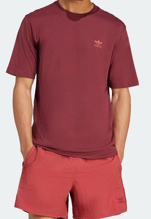 Homme portant un t-shirt Adidas bordeaux et un short Adidas rouge, mains dans les poches, visible du cou jusqu'au milieu de la cuisse sur fond uni.