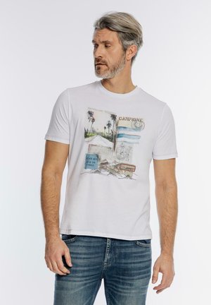 Mężczyzna z siwymi włosami i brodą, ubrany w biały t-shirt z nadrukiem i niebieskie dżinsy, patrzący w lewo na jednolitym tle.