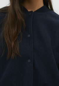 Marineblauwe wolmixjas met een ronde kraag, voorzien van zwarte knopen en een textuurafwerking. Close-up van de ontwerpdetails aan de voorkant.