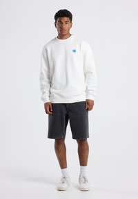 Valkoinen huppari, jossa on pieni sininen kukka-embroidointi, yhdistettynä harmaisiin farkkushortseihin ja valkoisiin tennareihin, joissa on hienovaraisia yksityiskohtia.