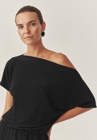Top negru off-shoulder dintr-un material texturat cu un ușor strălucire. Croi lejer, mâneci scurte și design drapat.