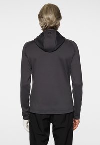 Uomo con giacca con cappuccio grigio scuro e pantaloni neri, visto da dietro su uno sfondo bianco semplice.