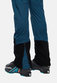 Blauwe en zwarte geïsoleerde skibroeken met zijritsen, gecombineerd met zwarte skischoenen met gekleurde accenten en geprofileerde zolen.