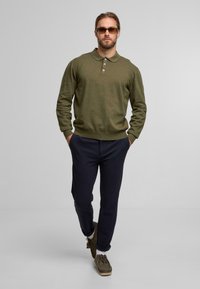 Man walking wearing an olive green long-sleeve polo, dark navy pants, brown shoes, white socks, and sunglasses against a plain background.