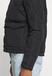 Černá prošívaná bunda s diamantovým vzorem, vybavená boční kapsou, manžetovými rukávy a lehkým, polstrovaným materiálem.