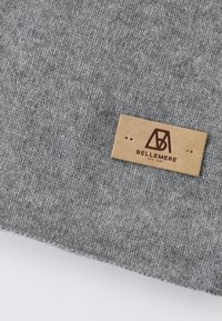 Tessuto lavorato a maglia grigio con una consistenza morbida, dotato di un'etichetta in pelle beige impressa con "BELLEMERE NEW YORK" e un logo geometrico.