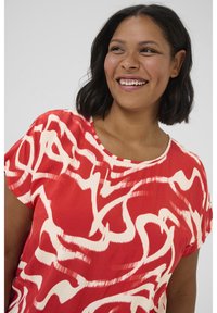 Haut rouge à manches courtes en tissu léger, avec un motif abstrait de coups de pinceau blancs et un col rond.