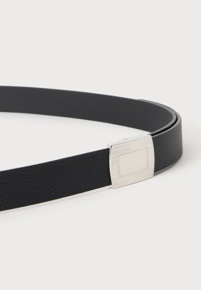 Ceinture en cuir noir texturé avec une boucle rectangulaire en métal argenté placée sur un fond uni clair.