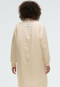 Langmantel in hellrosa mit gerippten Bündchen und Kragen, ausgestattet mit einem Patch-Aufnäher auf der Rückseite. Glattes Außenmaterial mit lässiger Passform.