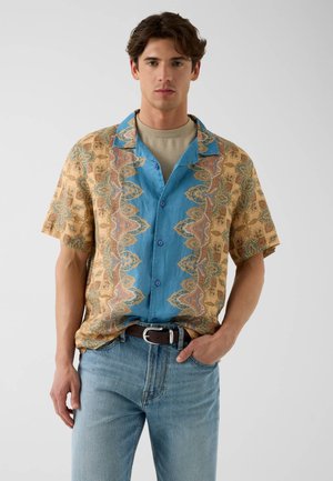 Man standing, wearing a patterned short-sleeve shirt with blue and tan sections, light jeans, and a brown belt, one hand in pocket.