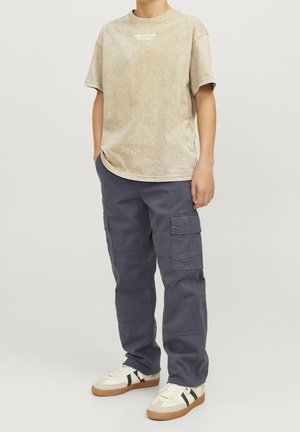 Camiseta beige de manga corta con textura desgastada y pequeño logo blanco; combinada con pantalones cargo oscuros con múltiples bolsillos y un ajuste relajado.