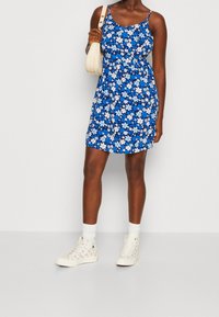 Kvinna i en blå blommig ärmlös klänning, vita högstövlade sneakers med vita strumpor, och bär en vit axelväska mot en ljus bakgrund.