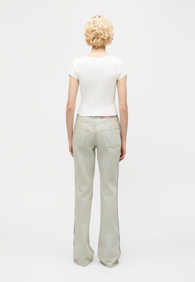 Άτομο με σγουρά ξανθά μαλλιά, φοράει εφαρμοστό λευκό κοντομάνικο μπλουζάκι και ανοιχτογκρίζα παντελόνια ίσιας γραμμής, στέκεται με την πλάτη προς τα εμάς.