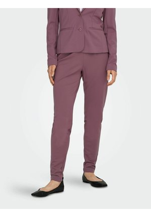 Vrouw draagt mauve aansluitende broek en bijpassende blazer, staand, gecombineerd met zwarte puntige platte schoenen op effen achtergrond.