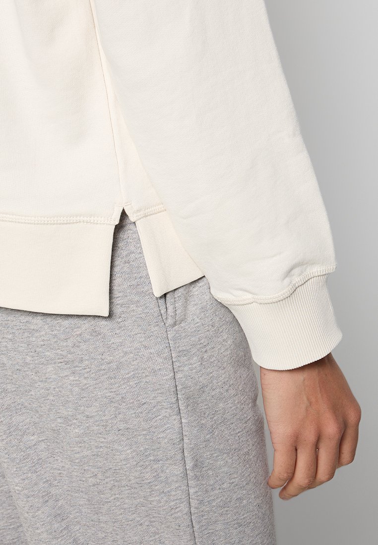 Felpa a maniche lunghe di colore crema con polsini a coste, abbinata a pantaloni della tuta grigi chiari. Presenta uno spacco laterale all'orlo per un comfort aggiuntivo.