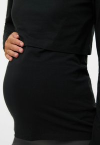 Crna majica s dugim rukavima od mekog materijala, s višeslojnim dizajnom koji prilagođava uočljivom okruglom trbuhu. Jednostavan i priljepljen siluet.