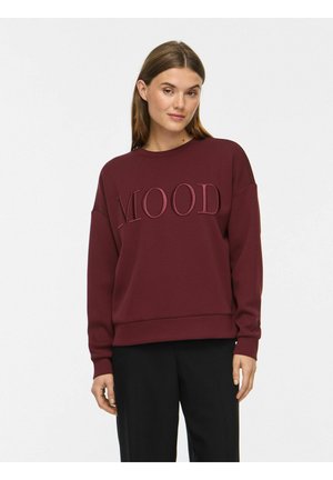 Jeune femme aux cheveux bruns et lisses portant un sweat bordeaux avec "MOOD" embossé et un pantalon noir sur un fond uni.