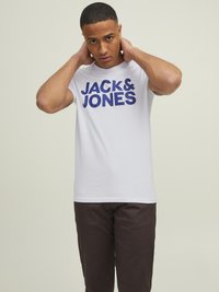 Bijela pamučna majica s hrabrim plavim printom "JACK & JONES" na prednjoj strani, u kombinaciji s tamnosmešnim hlačama. Ima okrugli izrez i kratke rukave.