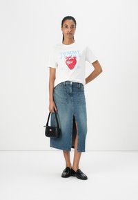 T-shirt blanc avec un motif de fraise rouge et un texte bleu, associé à une jupe en denim bleue taille haute avec une fente à l'avant. Sac à main et chaussures noires.