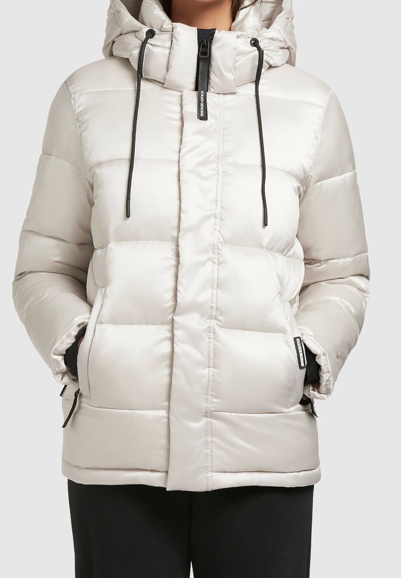 Chaqueta acolchada gris claro con capucha, que cuenta con una cremallera frontal, cordones negros y bolsillos laterales con cremalleras. Textura lisa y brillante.