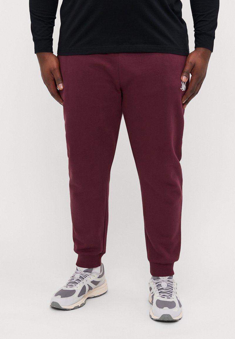 Pantalones deportivos burdeos con puños acanalados, hechos de una tela suave. Un pequeño logotipo es visible cerca del bolsillo. Se llevan con zapatillas grises.