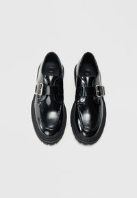 Pantofi din piele lacuită neagră, prevăzuți cu o curea cu cataramă, formă rotunjită, talpă groasă din cauciuc și detalii subtile de cusături. Textură netedă pe toată suprafața.