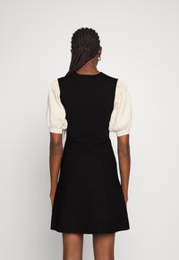 Robe noire avec un corsage ajusté et une jupe évasée. Manches bouffantes en tissu côtelé crème. Détail encolure en dentelle ajoute de la texture.