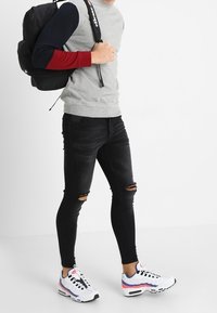 Maglione grigio con maniche rosse e blu, jeans neri strappati e scarpe da ginnastica bianche con dettagli rossi e blu. Zaino nero sulla spalla.