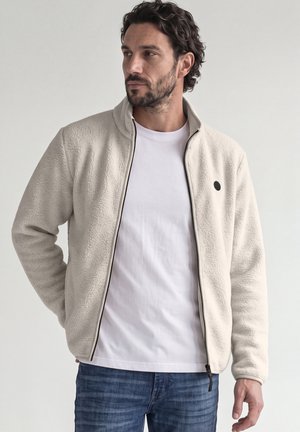 Homme en veste polaire blanche et t-shirt avec un jean bleu, debout devant un fond uni, regardant sur le côté.