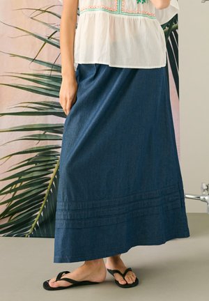 Kobieta ubrana w bezrękawnikową, haftowaną białą bluzkę, długą ciemnoniebieską dżinsową spódnicę i czarne klapki, stoi wewnątrz w pobliżu zielonych liści.