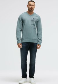 Hellblauer gestrickter V-Ausschnitt-Pullover mit "CAMP DAVID"-Schriftzug, kombiniert mit dunkler Jeans und weißen Sneakers. Eng anliegender Stil und gerippte Bündchen.