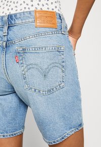 Vaaleansiniset farkkushortsit, joissa on klassinen viiden taskun malli, nahkakangas, jossa on brändimerkintä, ja erottuva kaareva takataskujen ommel.