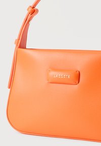 Πορτοκαλί δερμάτινη τσάντα ώμου με ρυθμιζόμενο λουράκι και λογότυπο Lacoste με ανάγλυφα χρυσά γράμματα στο κέντρο μπροστά.